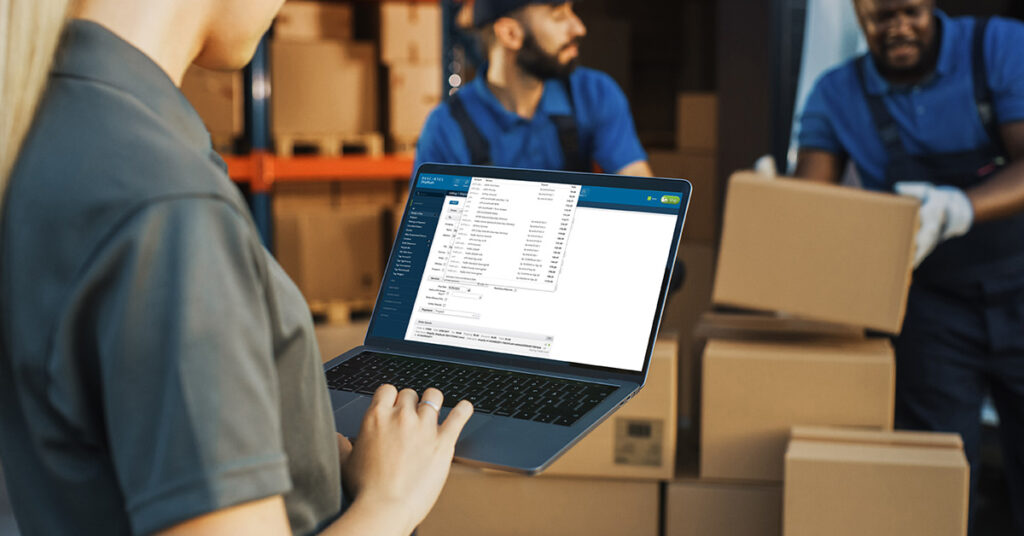 Warehouse manager holding a laptop computer displaying Descartes ShipRush shipping software, use as ShipStation alternative to print shipping labels in a busy warehouse with boxes and warehouse workers