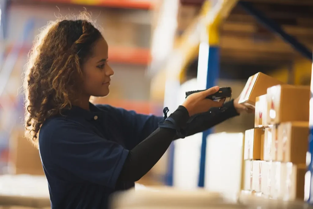 A female warehouse worker using a warehouse management system with mobile barcode scanning to scan a parcel on a shelf.