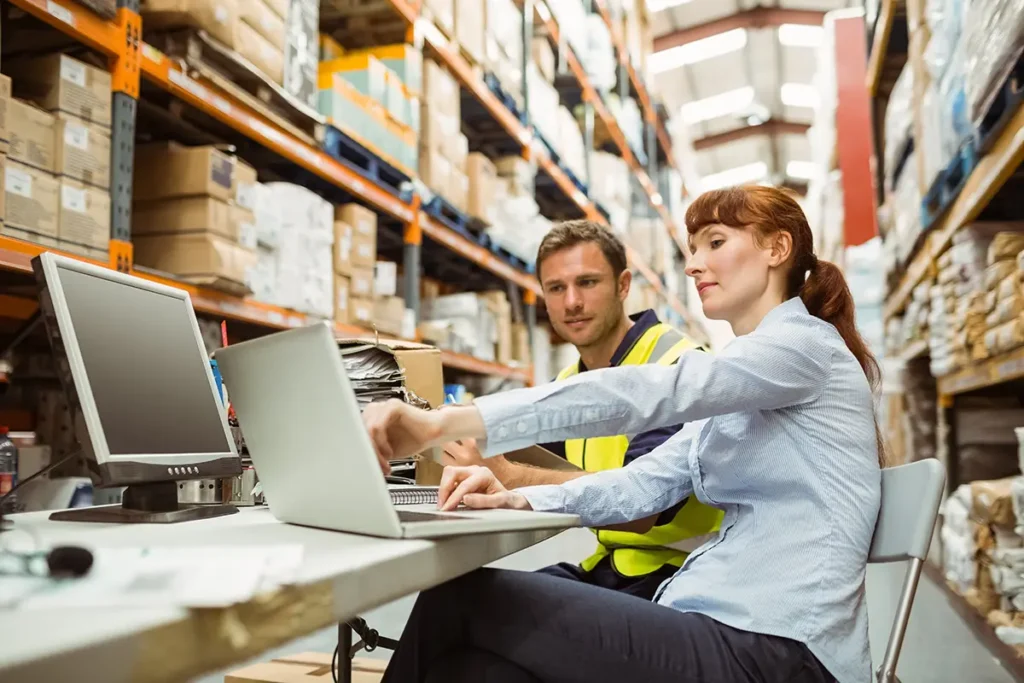 A manager and warehouse worker sitting together in front of a laptop computer in the warehouse as the manager points out how warehouse shipping software is reducing shipping costs.