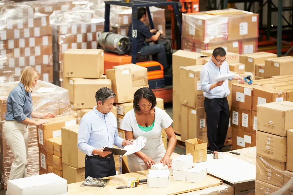 A busy warehouse with five people working in an area crowded with boxes, a forklift, and a shipping station without warehouse shipping software or computers.