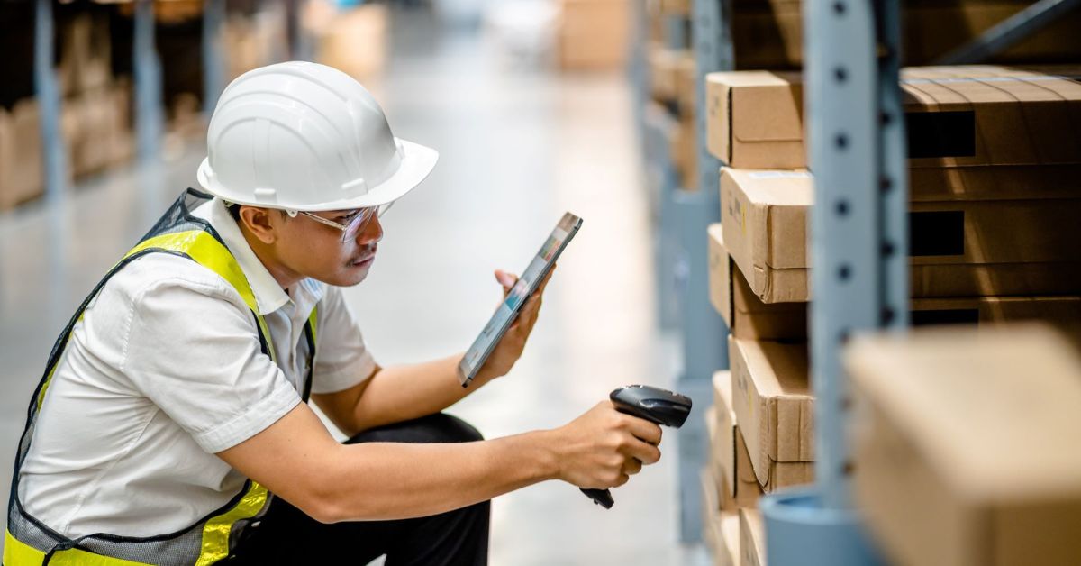 A warehouse worker holding a mobile device with a warehouse management system, using a barcode scanner to scan a box on a warehouse shelf.