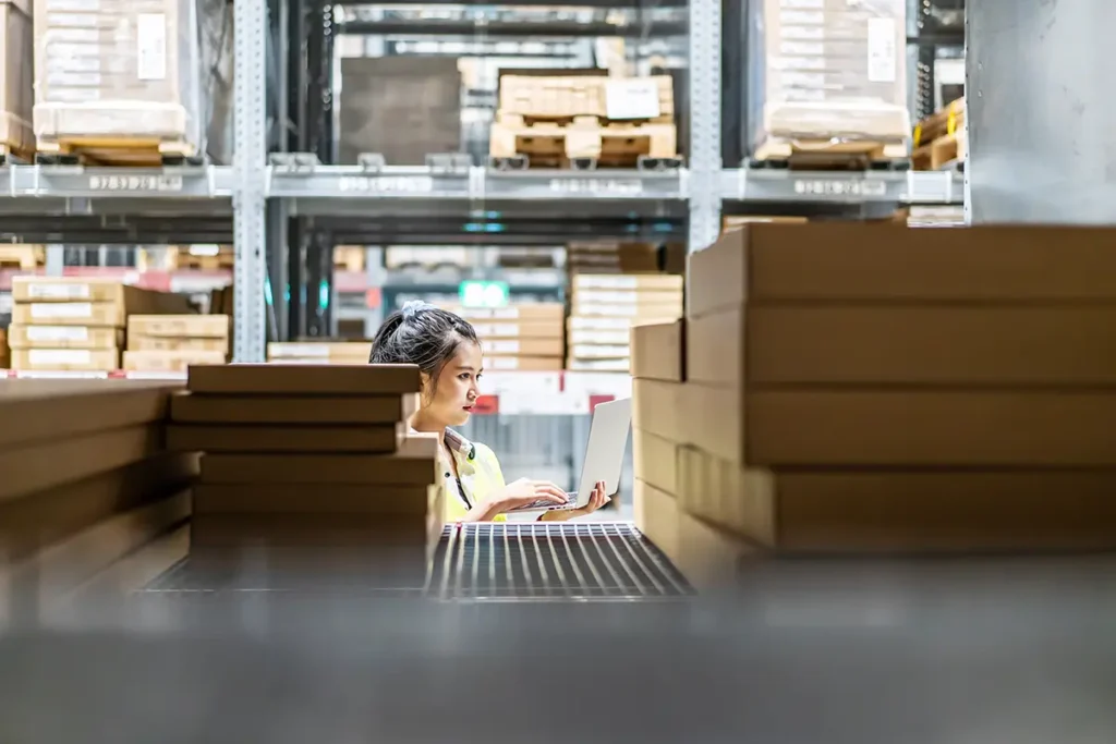 Alt text: Female warehouse worker surrounded by piles of boxes for ecommerce shipping, typing on a laptop using a manual process