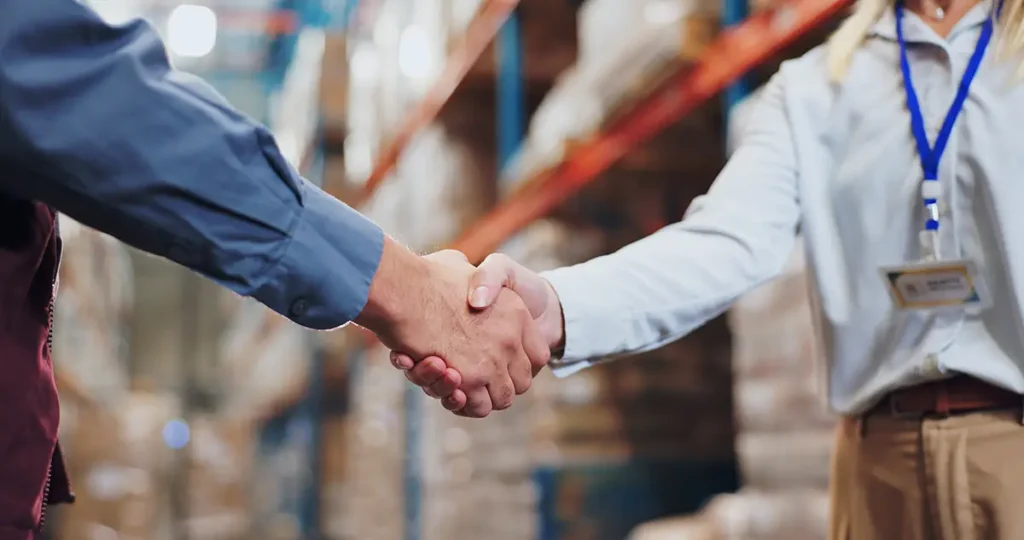 Shipping software provider and shipping carrier representative are partners shaking hands in an ecommerce warehouse.